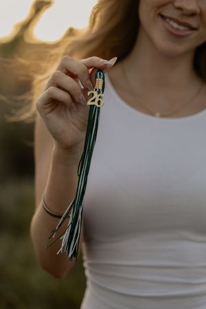 Smiling woman holding green and white graduation tassel with 26 charm at golden hour.