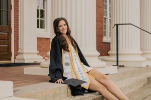 A smiling female graduate in black cap and gown sits on steps before white columns at her college graduation.