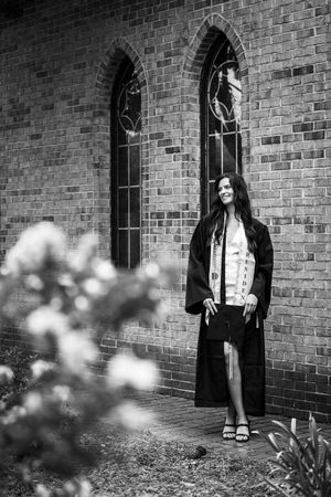 Black and white photo of a female graduate in cap and gown standing beside a brick church.