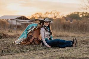 Cowgirl in turquoise fringe vest and hat sitting against western saddle at golden sunset.