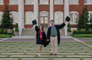 Two graduates in caps and gowns celebrating with raised mortarboards in front of brick university building.