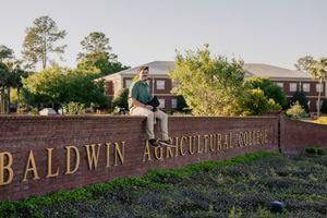 Male graduate sitting on Baldwin Agricultural College brick sign surrounded by lush green trees.