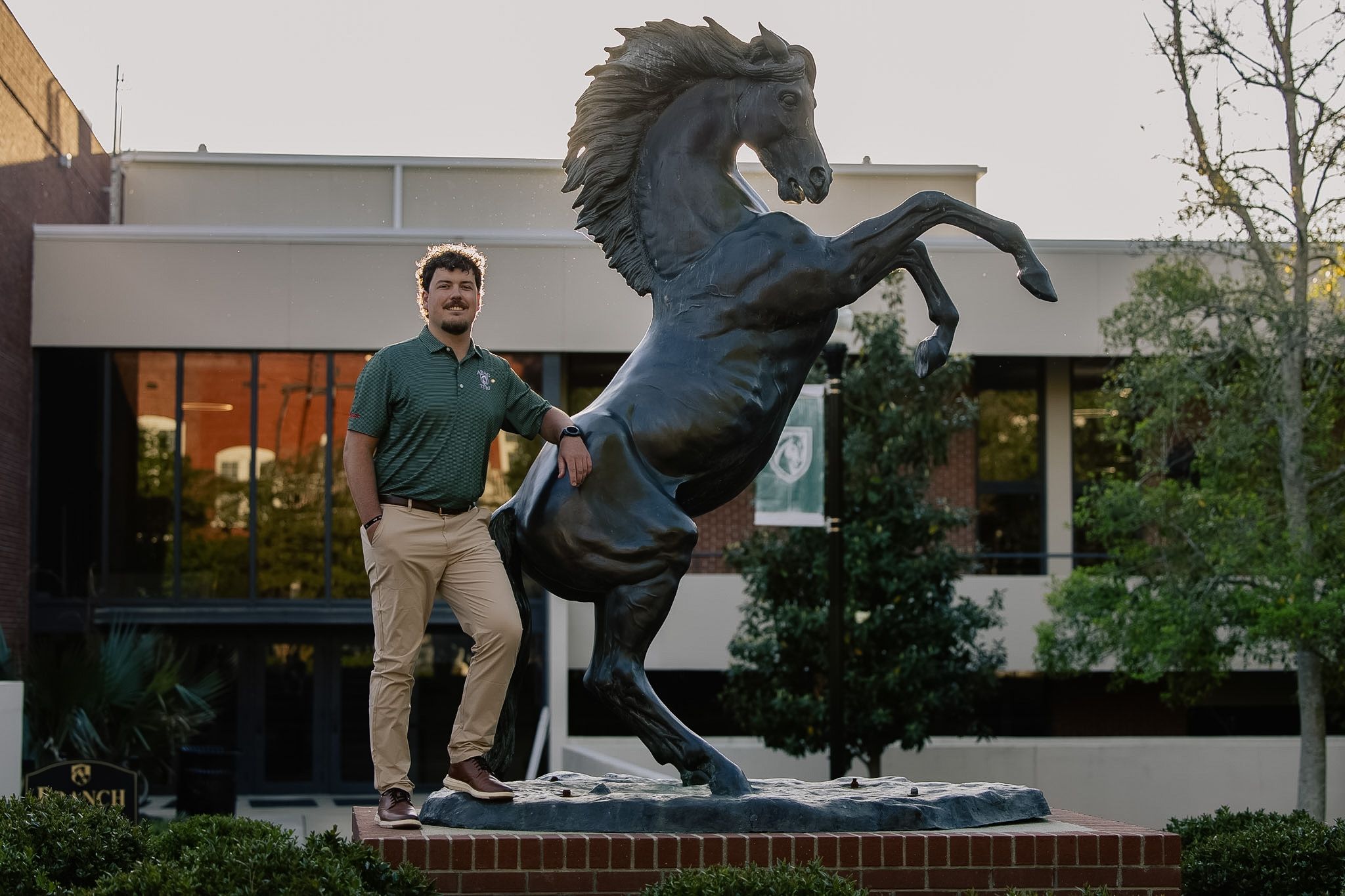 A young man stands proudly beside a rearing bronze mustang horse statue on a college campus.