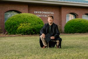 Male graduate kneeling on grass lawn in front of Environmental Horticulture brick building.