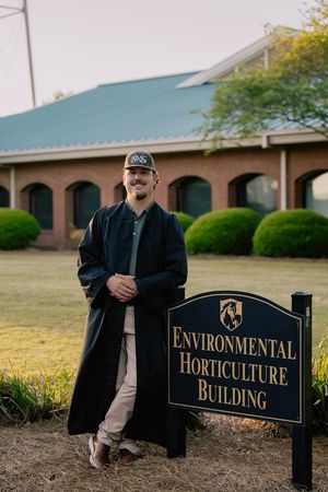 Male graduate in black gown standing beside Environmental Horticulture Building sign at brick campus.