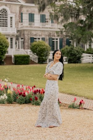 Woman in floral maxi dress standing on gravel path with historic stone mansion and flowers behind her.