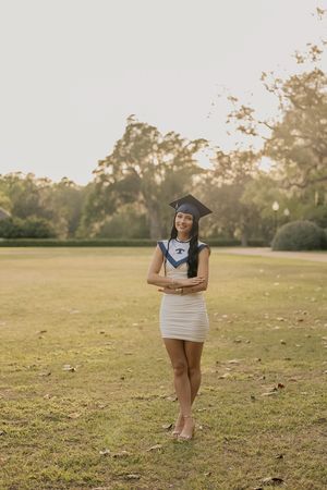 Female graduate in cap holding diploma in white dress standing on green lawn at golden hour.