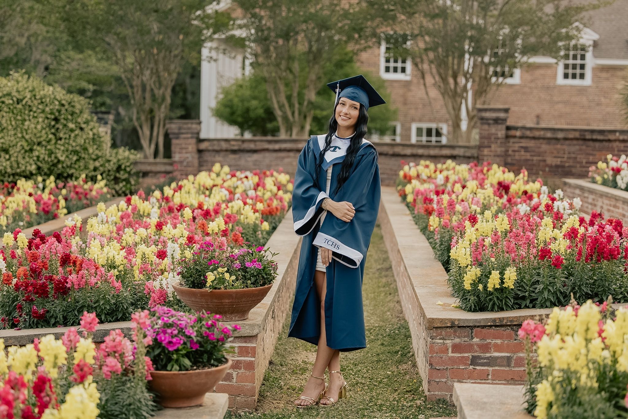 Female graduate in blue cap and gown posing among colorful spring flower gardens at brick building.