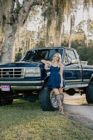 Blonde woman in blue dress and cowboy boots poses beside lifted Ford truck under Spanish moss trees.