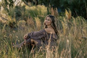 Young woman in brown off-shoulder dress sitting gracefully in golden tall grass during sunset.