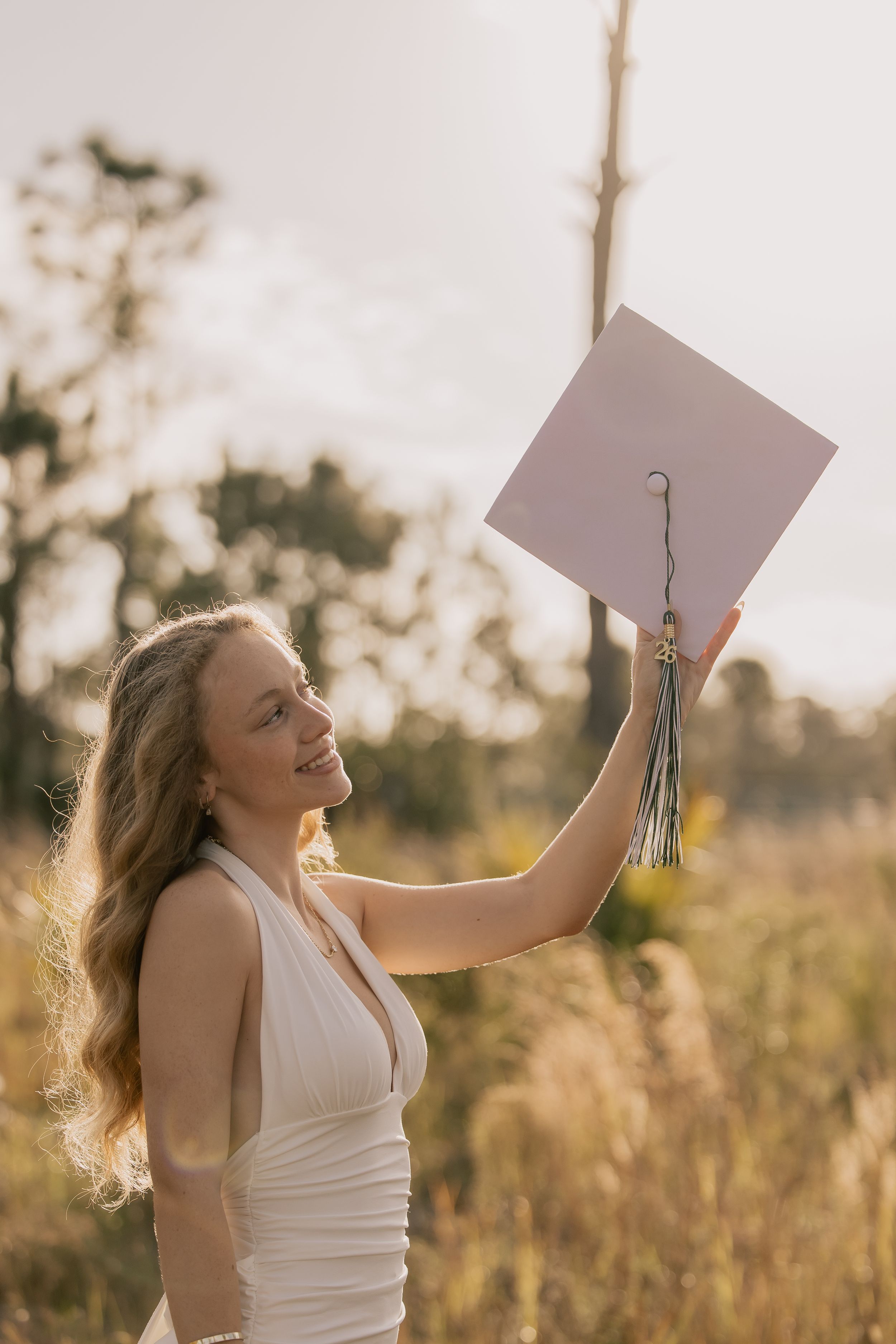 Smiling young woman in white dress holds graduation cap aloft in sunlit field during golden hour.
