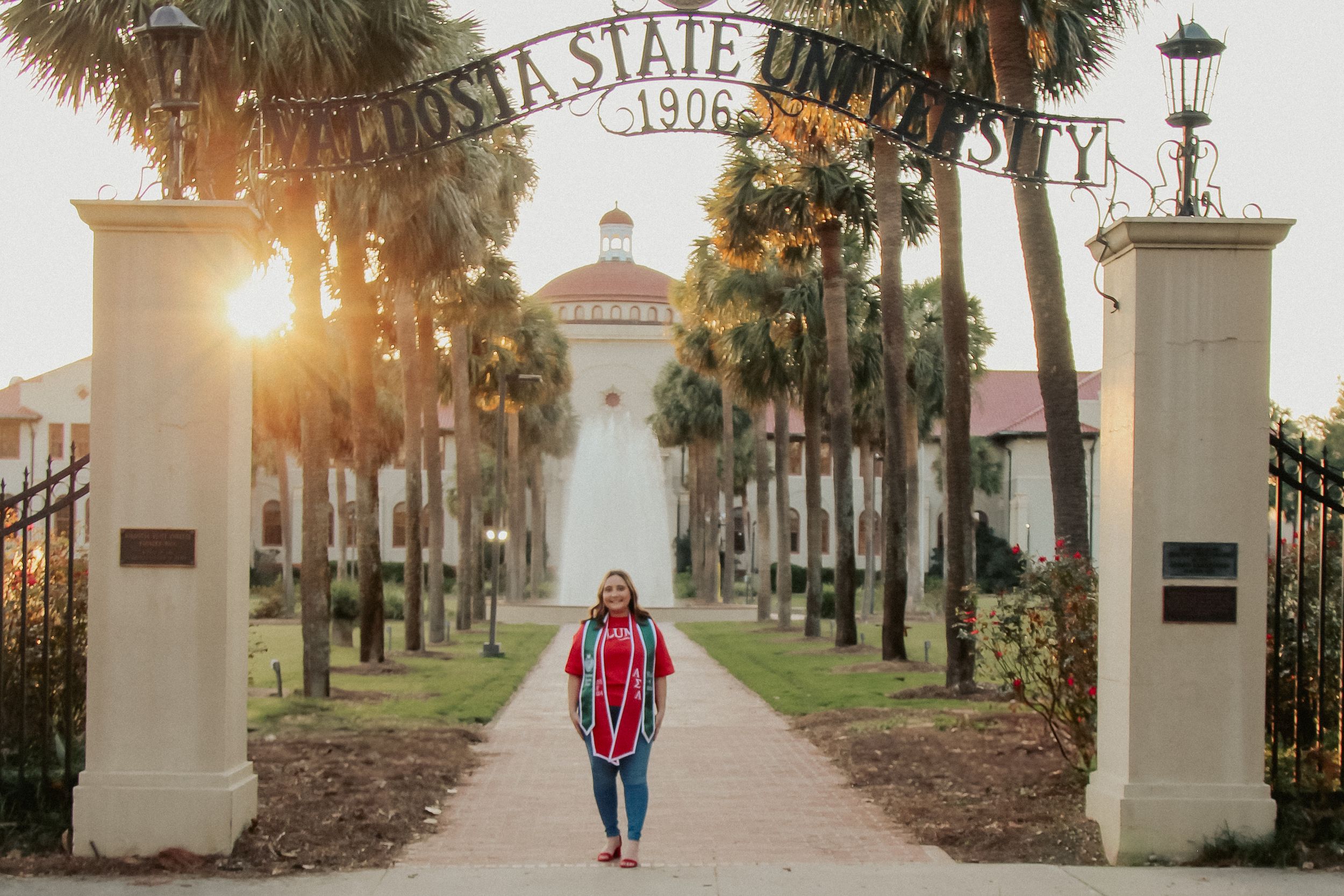Graduate standing at Valdosta State University entrance gate at golden sunset with palm trees.