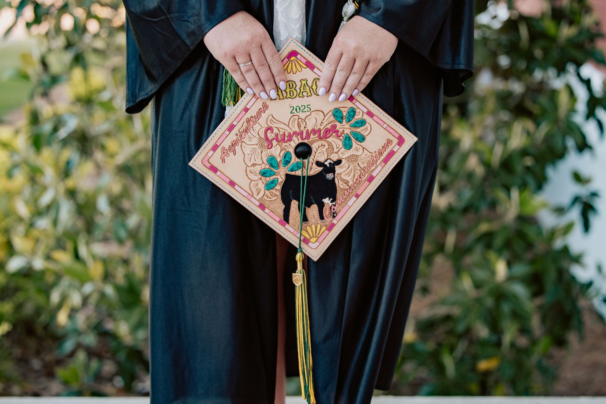 Graduate in black robe displays decorated graduation cap with colorful floral artwork and tassel.