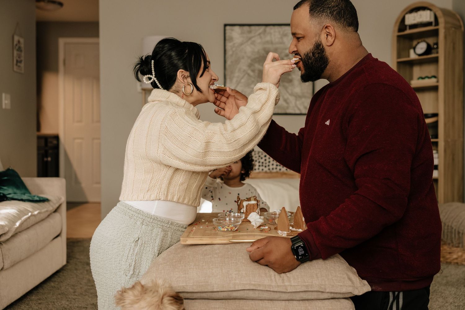Two people playfully interact while standing at a kitchen counter with a cozy home interior in the background.