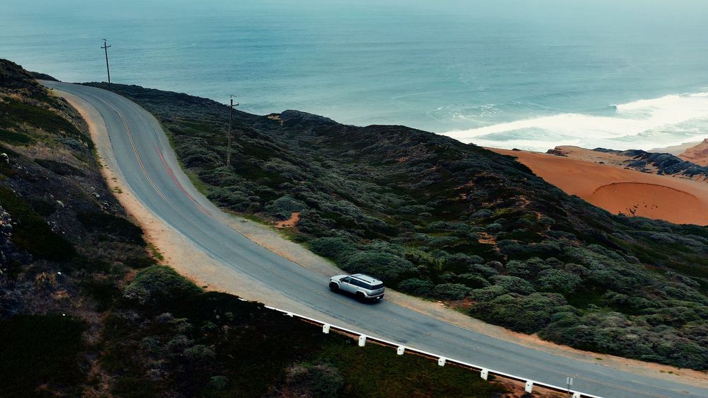 Coastal highway curves along Pacific shoreline with white vehicle driving against blue ocean.