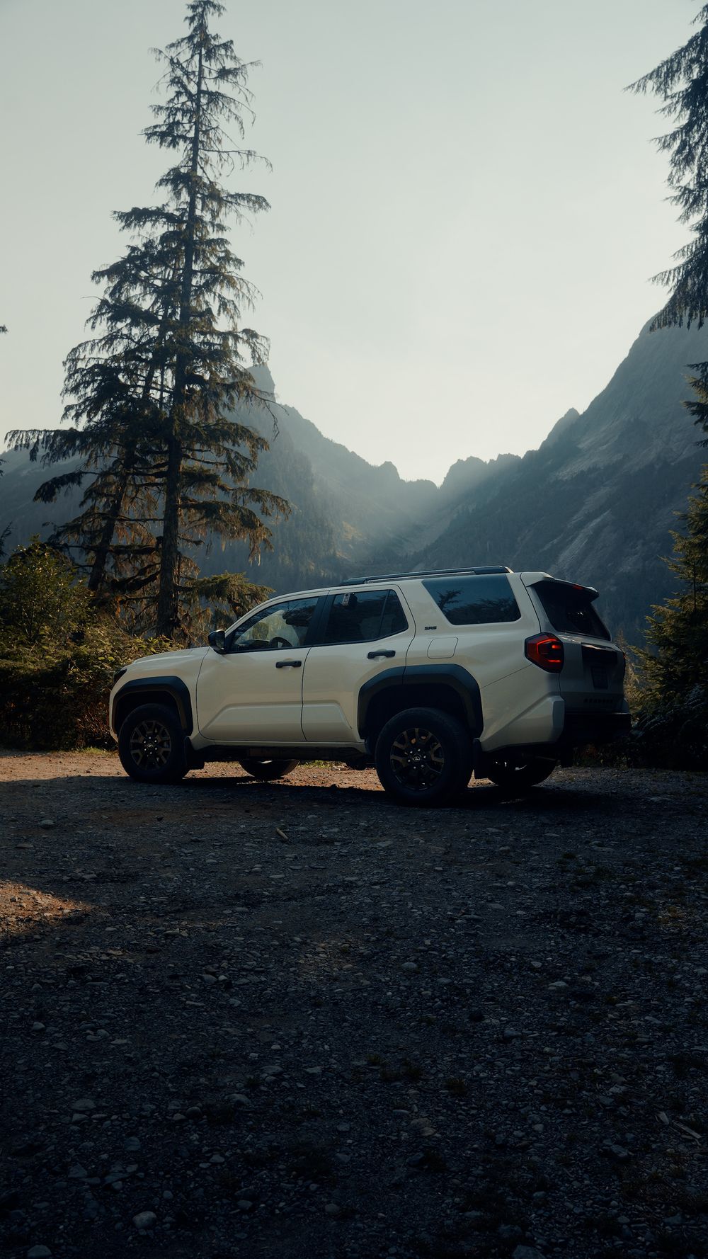 White SUV parked on rocky mountain overlook with evergreen trees and misty peaks in background.
