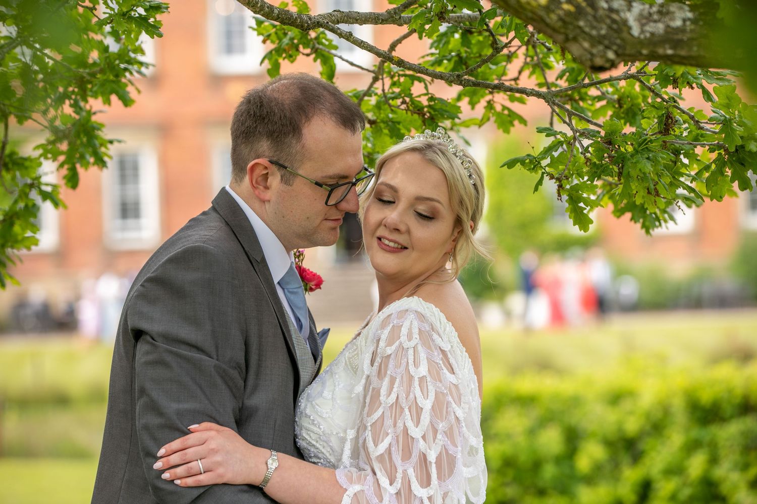 A couple embraces under a leafy tree in front of a brick building. The bride wears a lace gown, the groom a suit. The mood is serene