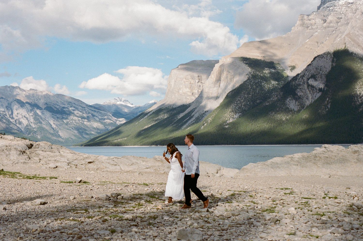35 mm film photo of bride and groom walking along the rocky shoreline of Lake Minnewanka in Banff with mountains in the background.