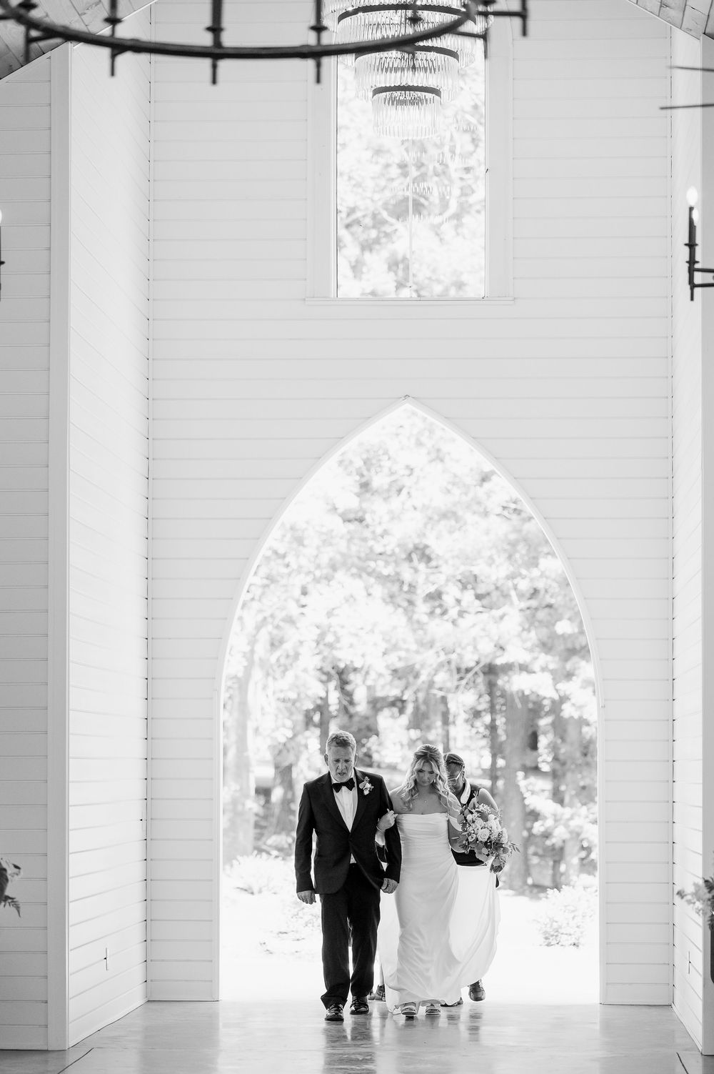 A black and white photo of a couple walking through a gothic arched doorway in a bright chapel.