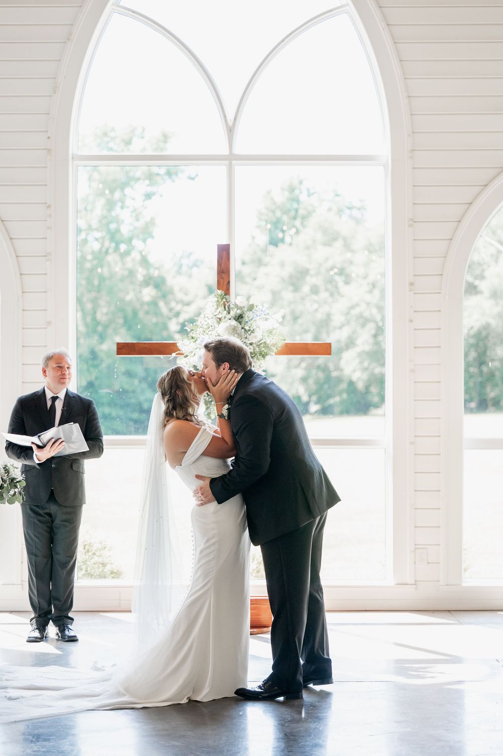A wedding ceremony unfolds in a bright chapel with arched windows and wooden cross.