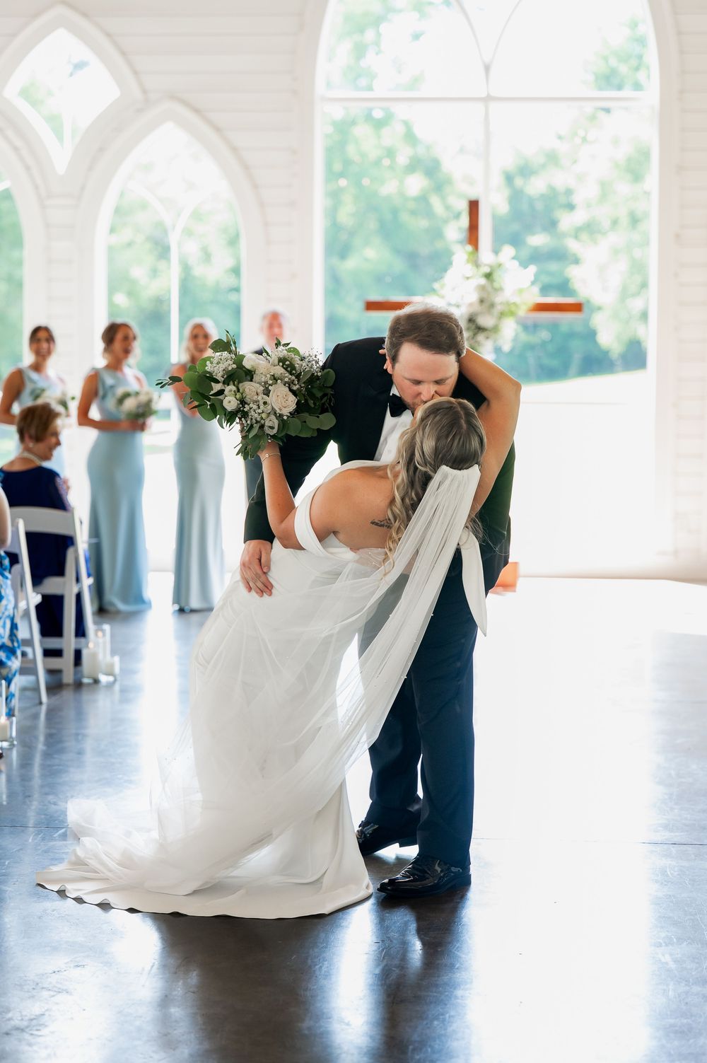 A romantic dip during a wedding dance in an elegant chapel with gothic arched windows and bridesmaids in blue dresses.