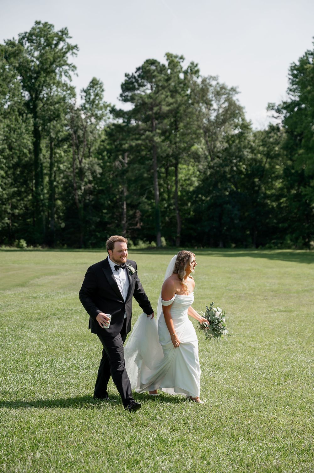 A wedding couple walks hand in hand across a sunny green lawn surrounded by tall trees.