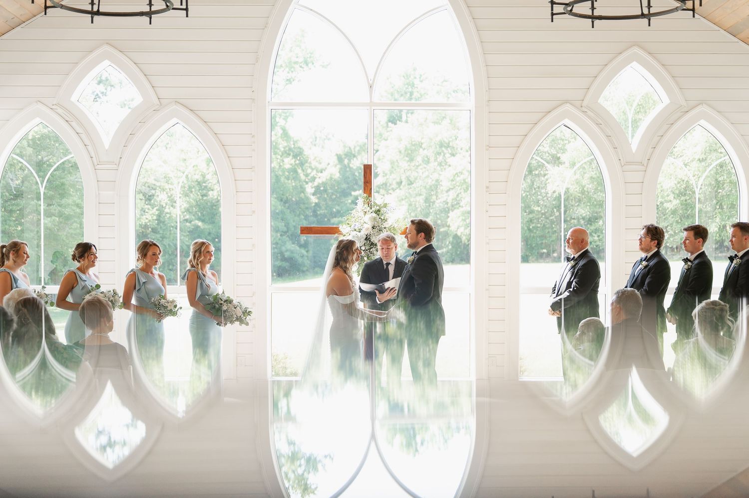 Wedding party members stand in a row against arched windows in an elegant white venue.