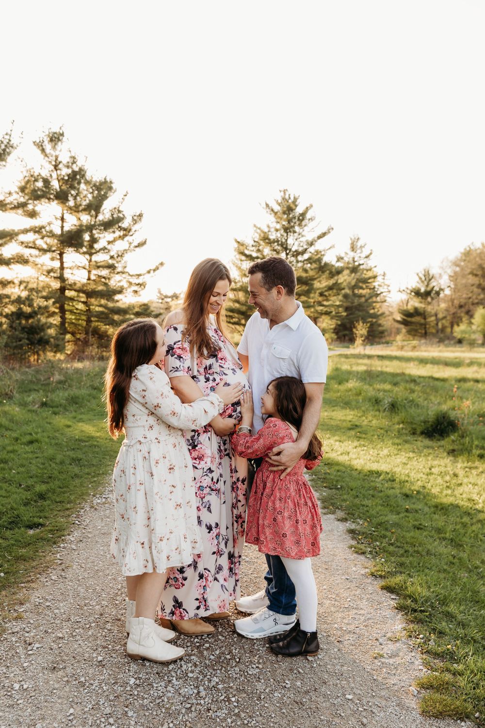 Family shares tender moment together on a sunlit dirt path surrounded by evergreen trees and grass at golden hour. cleveland maternity photo