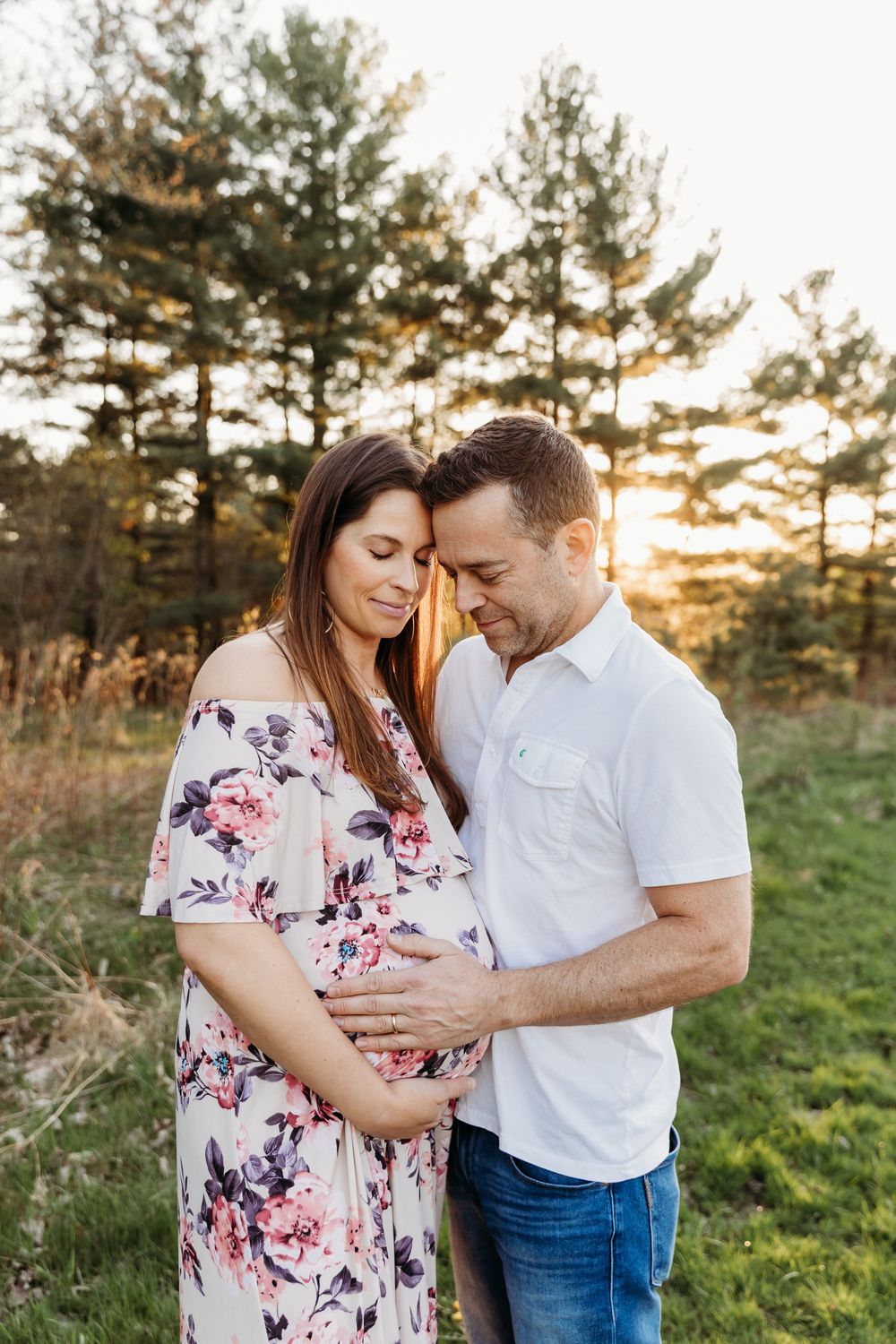 A couple shares an intimate moment in a sunlit field with pine trees during a maternity photoshoot at sunset.