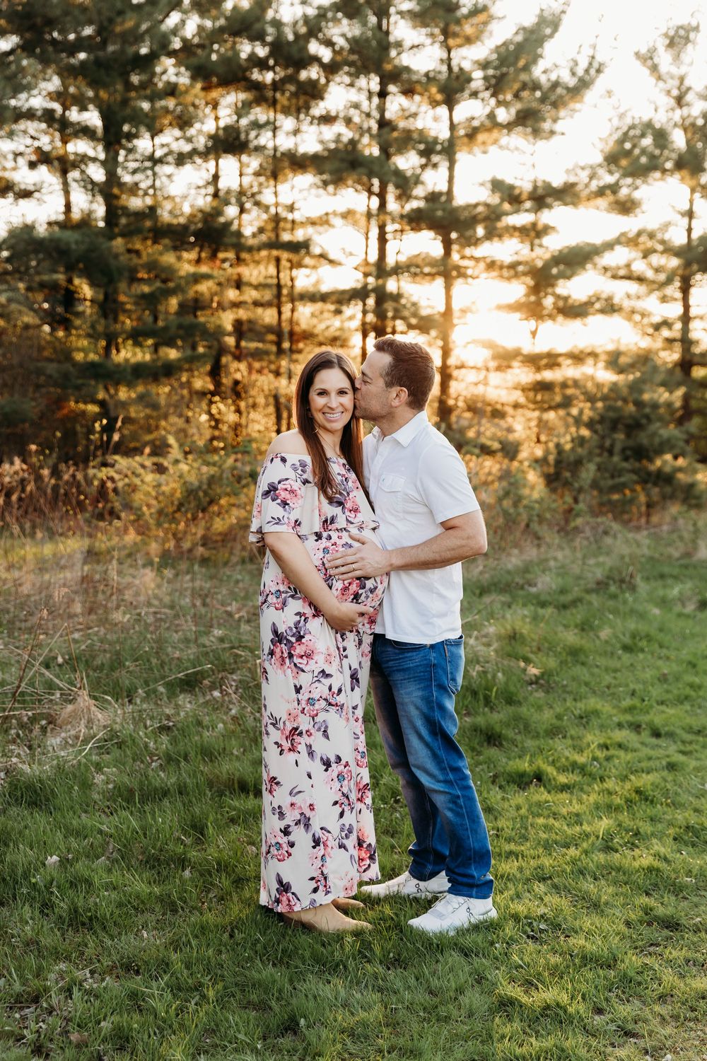 A couple shares tender moments at sunset in a grassy field with pine trees in the background.