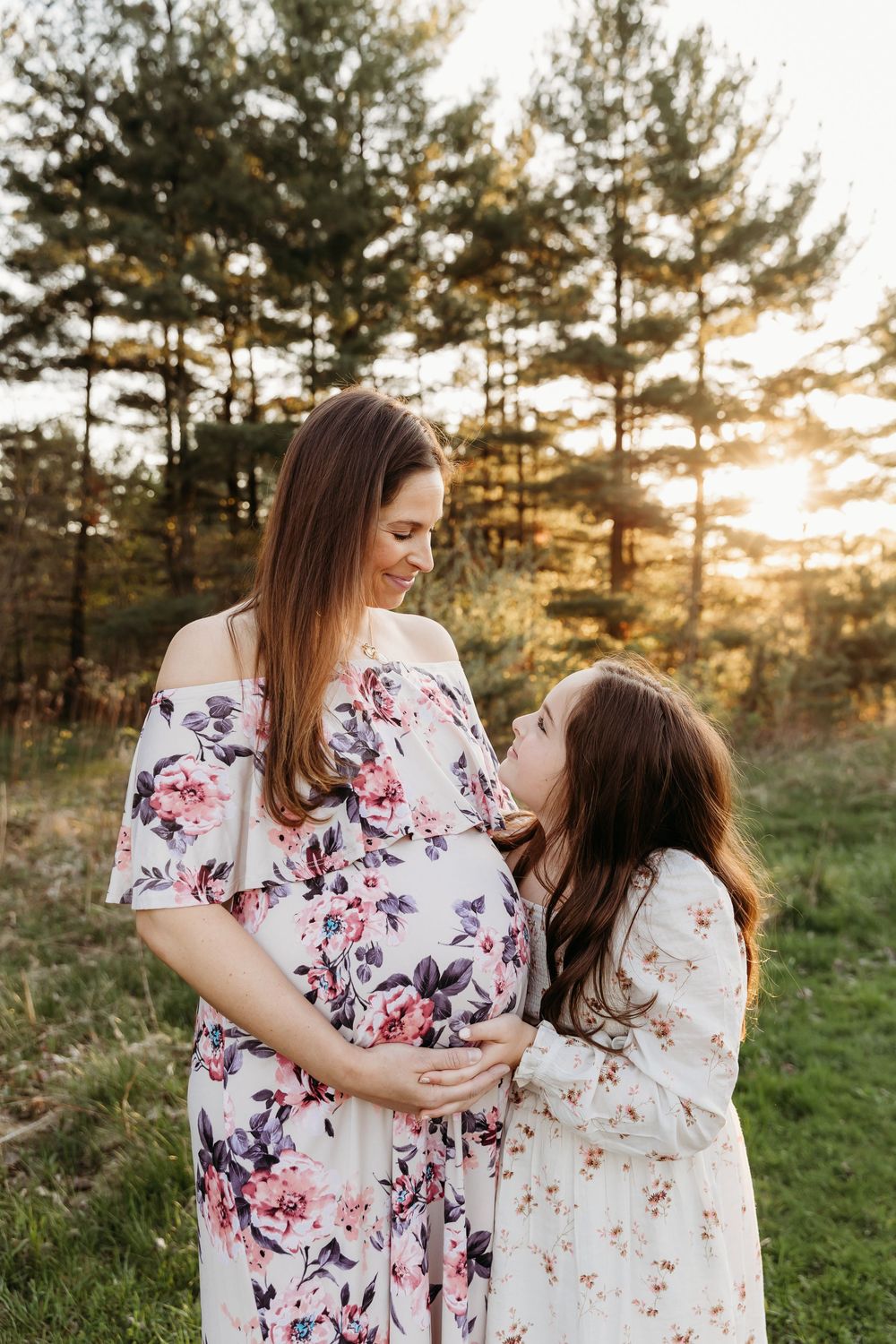Mother and daughter wearing floral dresses embrace during golden sunset in pine forest setting.