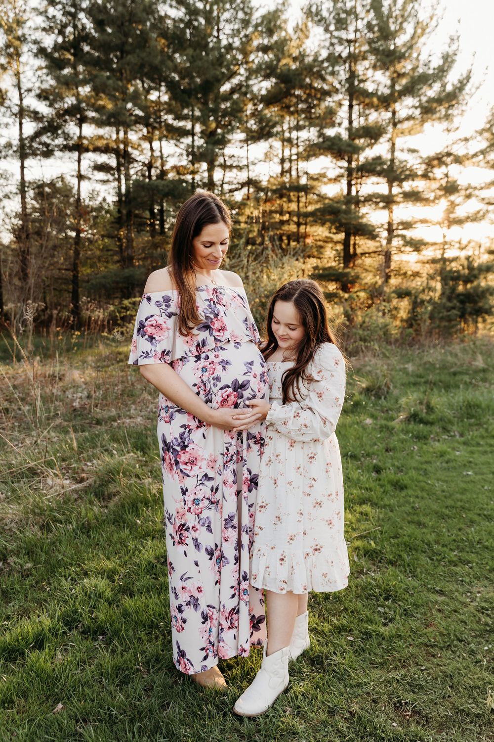 Two people in matching floral dresses embrace while standing in a grassy field at sunset with pine trees in background.