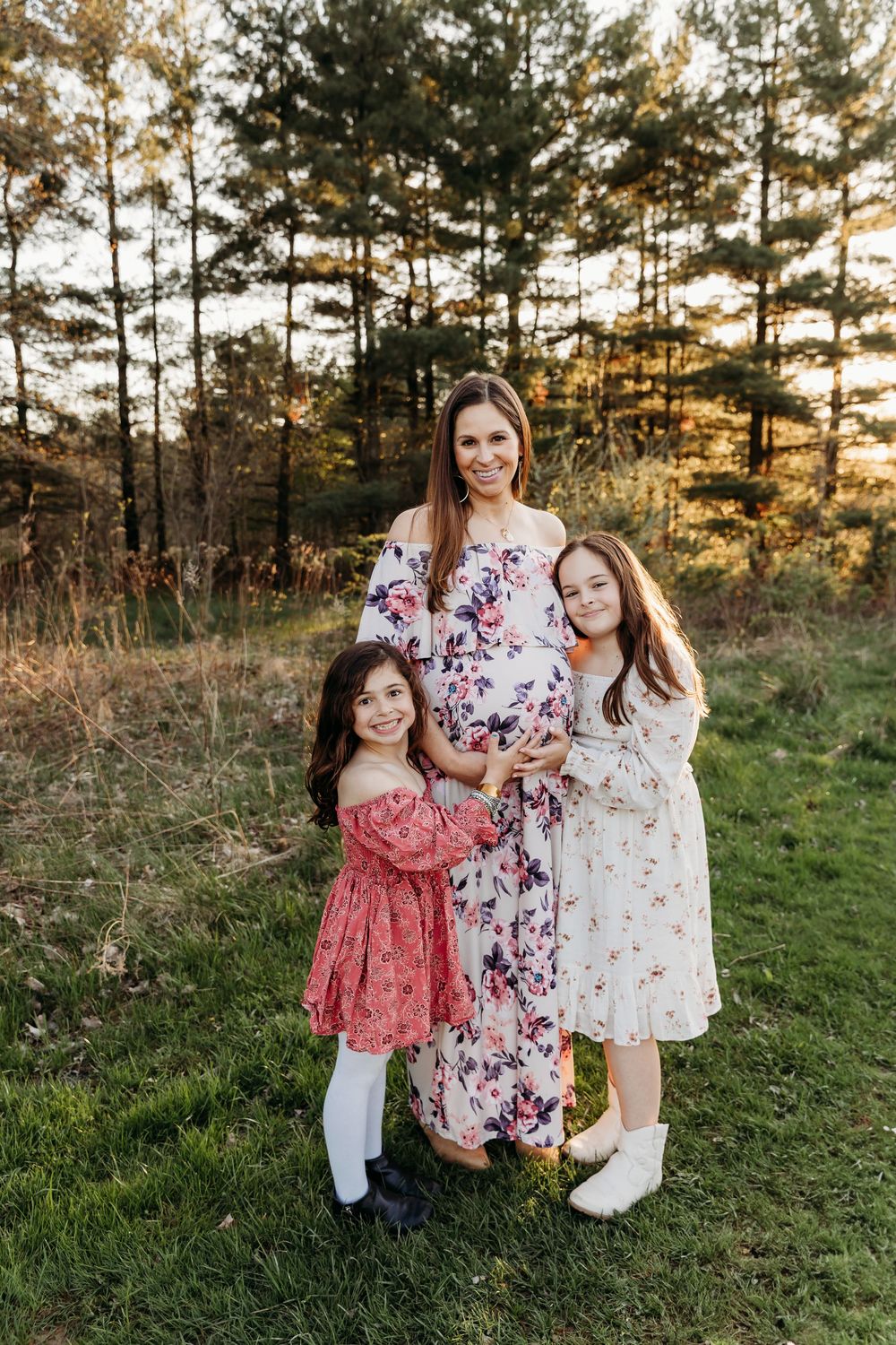 Family wearing floral dresses poses together in a grassy field at sunset with pine trees in the background.