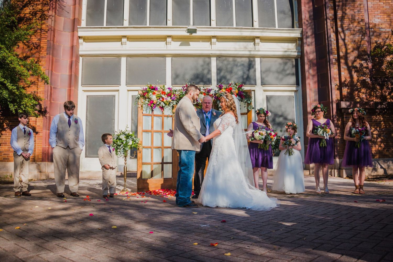 Larry and Misty hold hands at their floral altar during a ceremony at The Katy Depot in Denison, TX. Photography by Wolfpack Wedding Co.