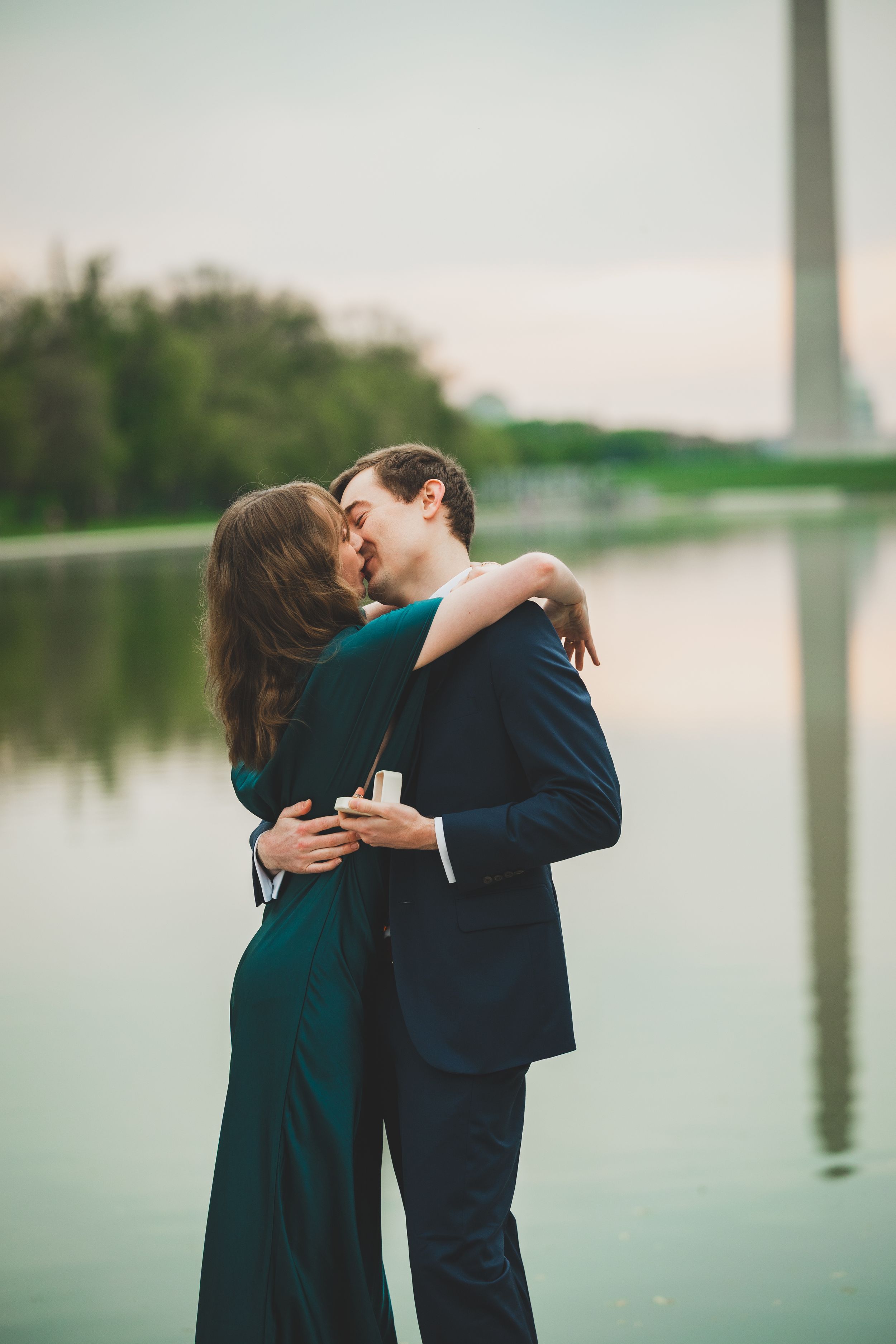 A romantic couple embraces and kisses near a reflective pool with the Washington Monument visible in the background.