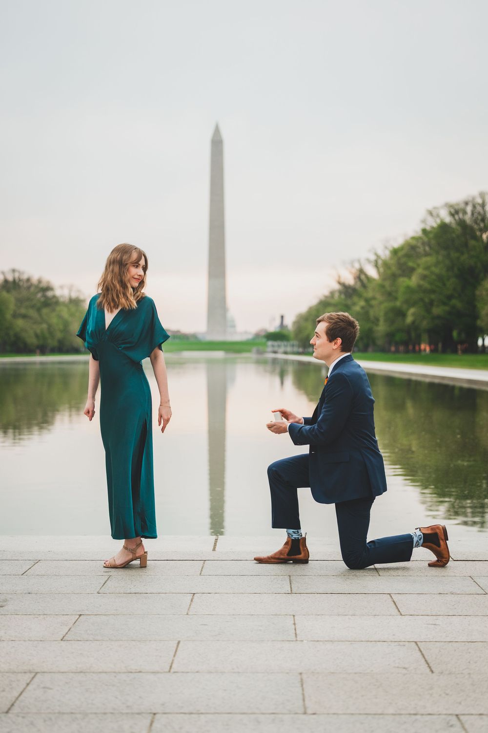 A romantic proposal scene unfolds by the reflecting pool at the Washington Monument during sunset.