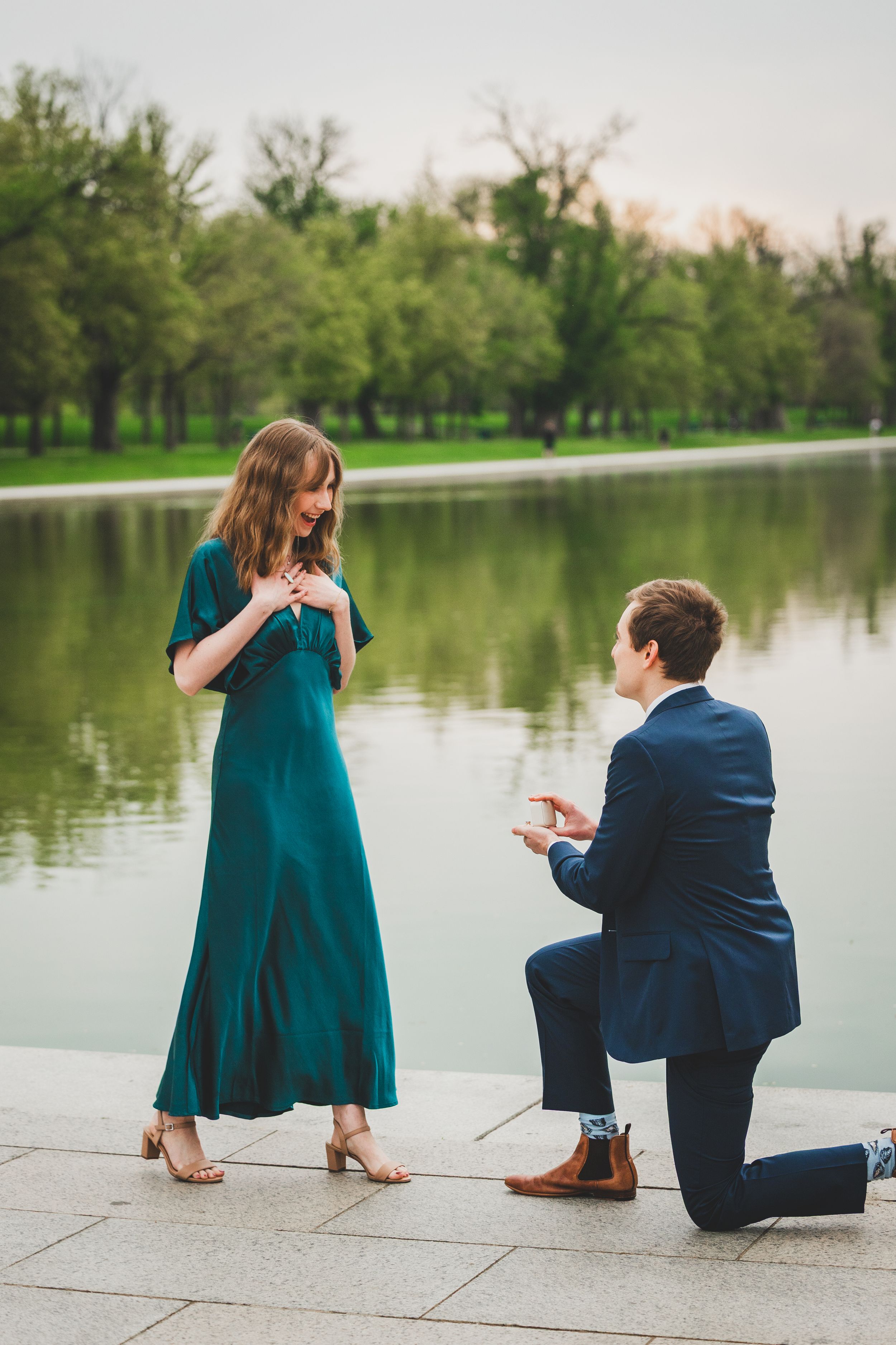 A couple sits by a serene lake at sunset, with the person in teal dress and another in navy suit.