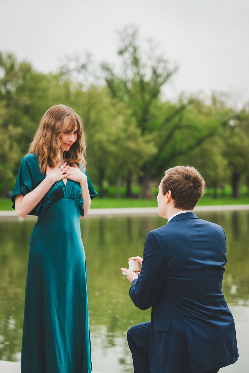 A romantic marriage proposal scene by a lake with a person in a teal dress and another in a navy suit kneeling.