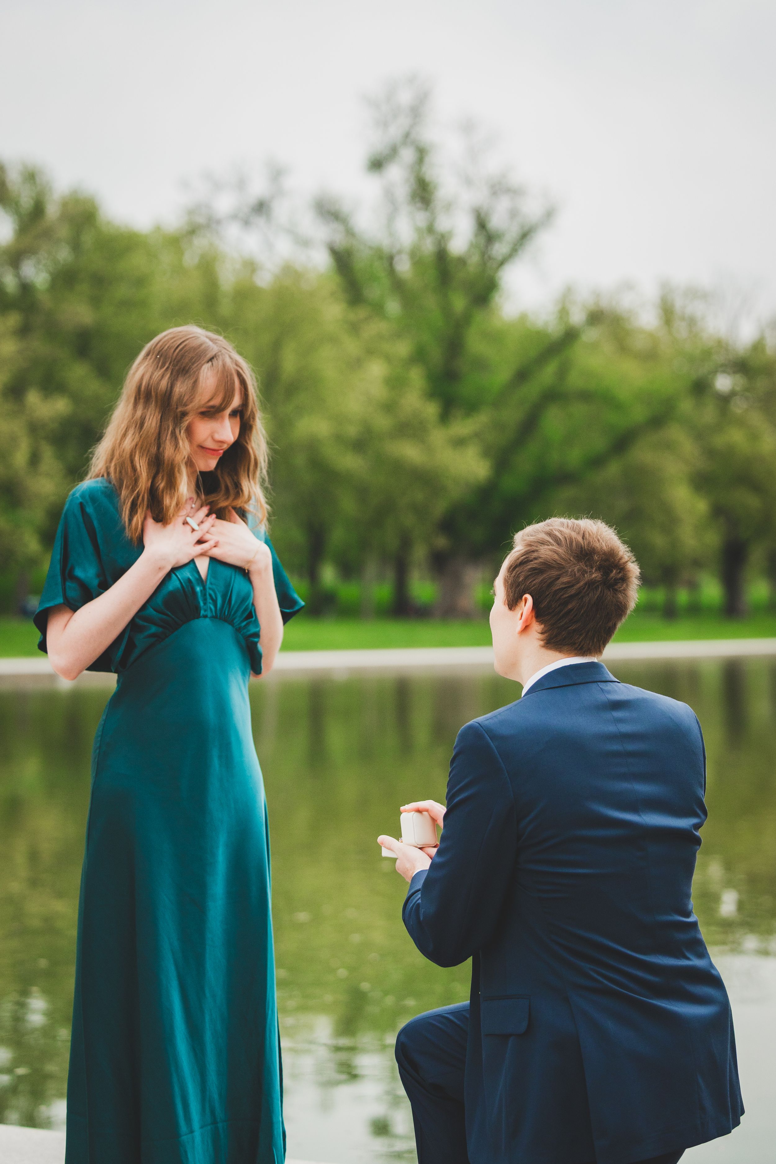 A romantic marriage proposal scene by a lake with a person in a teal dress and another in a navy suit kneeling.