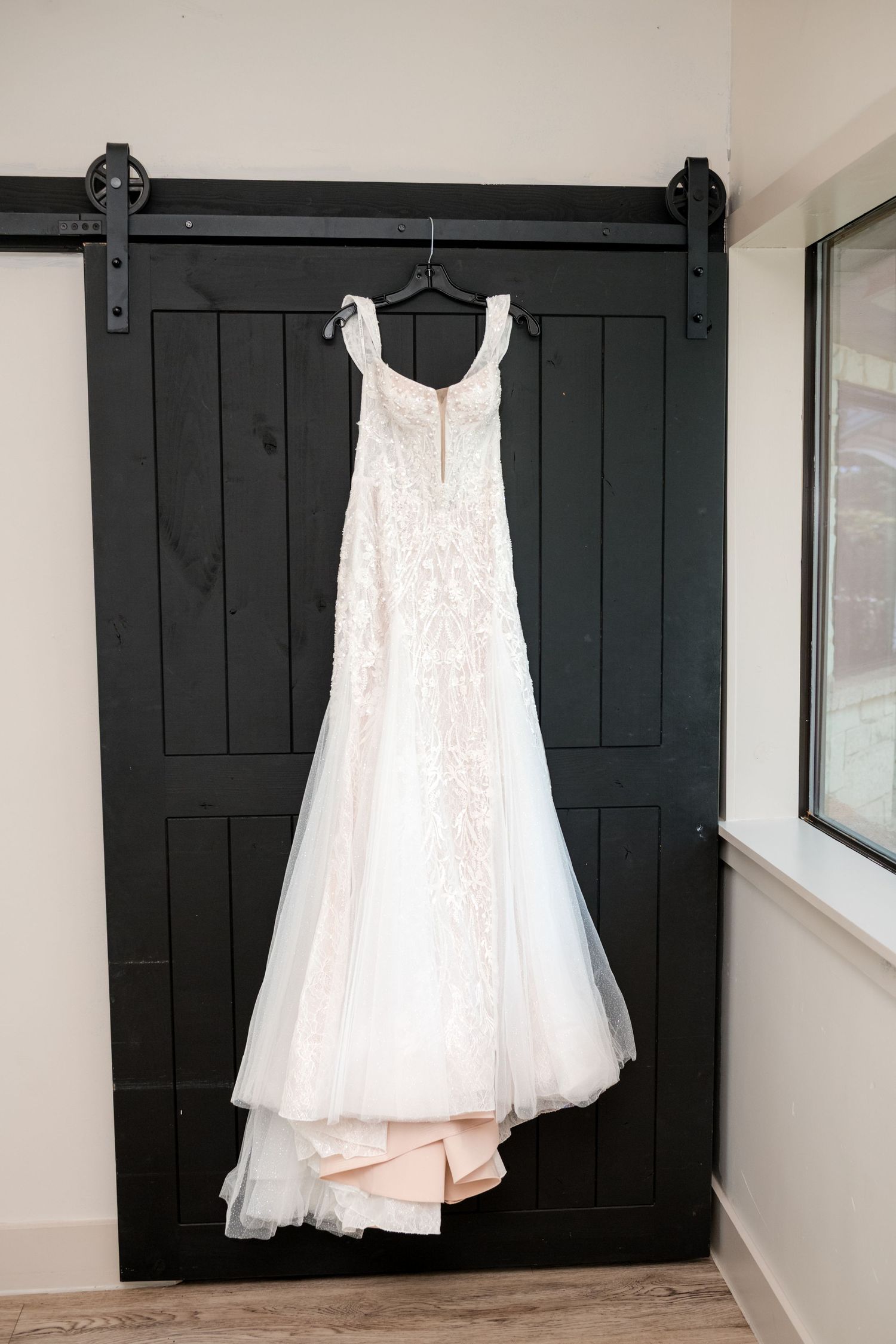 White lace wedding dress hanging on a black barn door in a bright interior hallway.