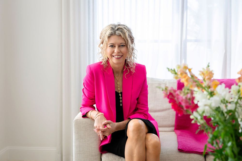 A person in a bright pink blazer sits at a desk with flowers and natural lighting from nearby windows.