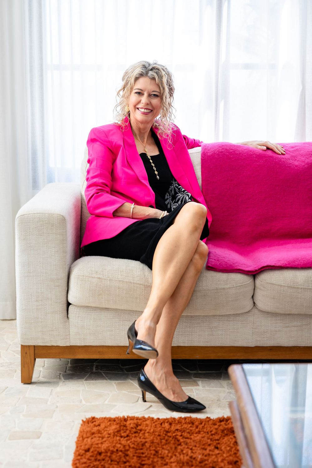 Professional woman in hot pink blazer and black dress sits elegantly on beige couch with pink accent pillow.