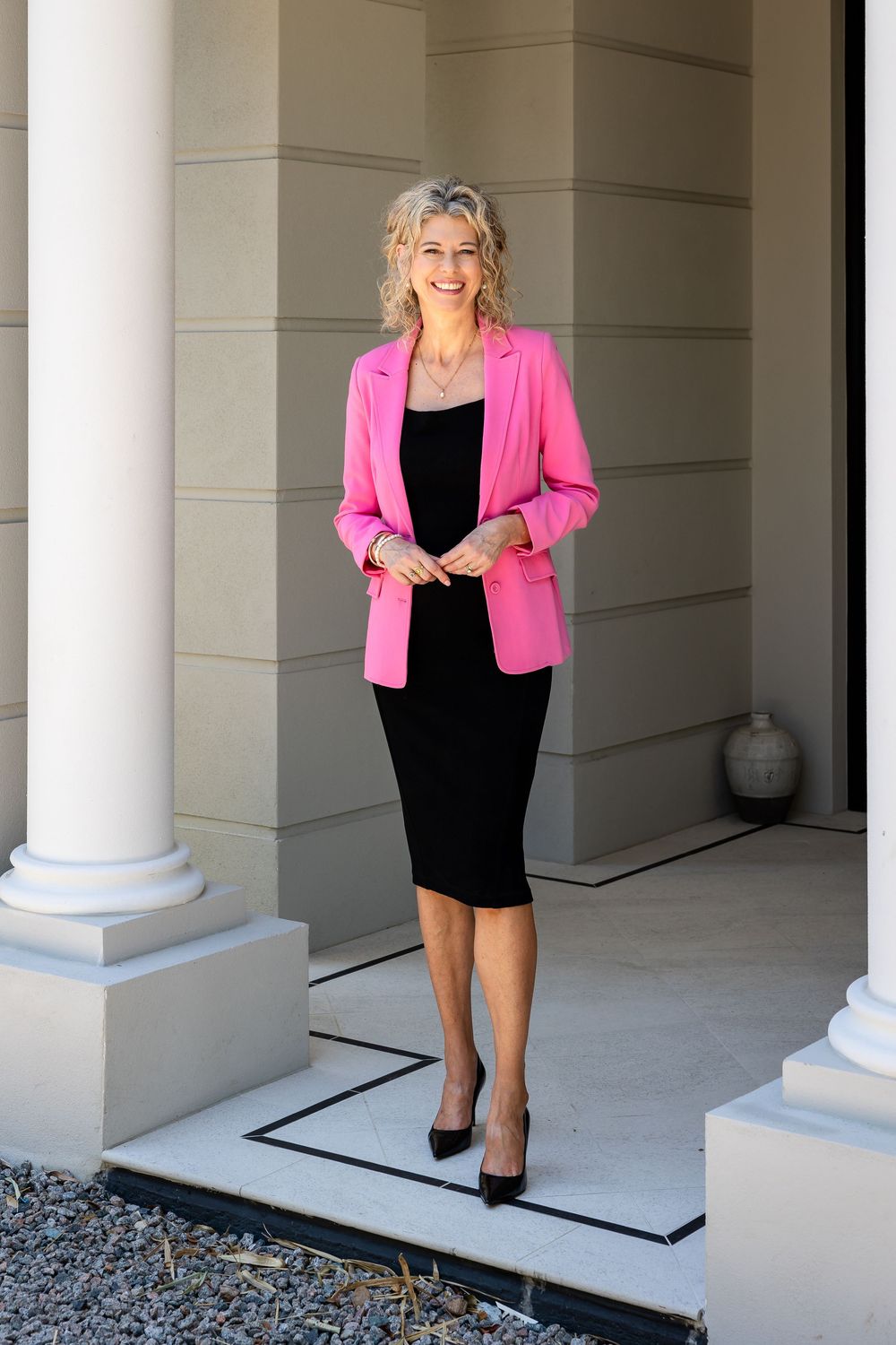 Professional woman in a bright pink blazer and black dress posing in a columned hallway.