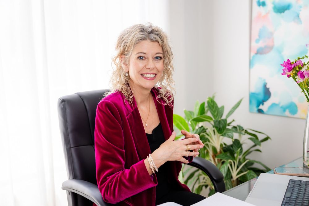 Professional in a burgundy velvet blazer sits at a desk with office plants and blue artwork in the background.
