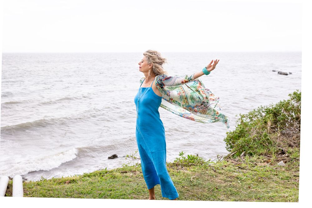 Woman in bright blue dress dances joyfully on grassy oceanfront cliff at sunset.