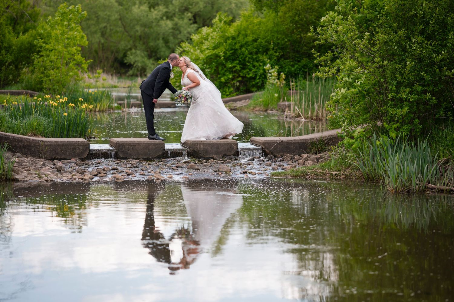A bride in a white wedding dress poses on a wooden bridge over a tranquil pond surrounded by lush greenery.
