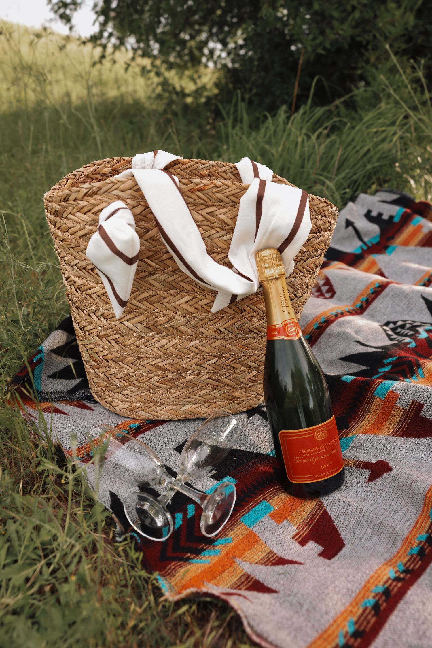 Straw beach bag and champagne bottle on colorful blanket with sunglasses set for outdoor picnic.