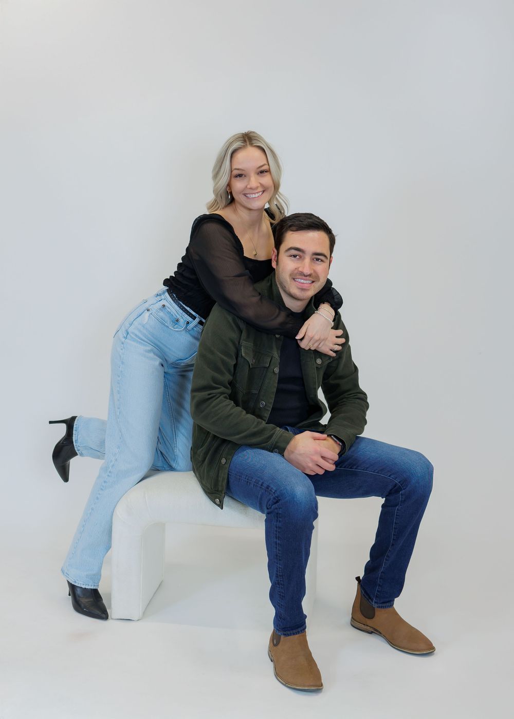 A couple poses together in a studio setting wearing casual outfits of jeans and jackets against a white backdrop.