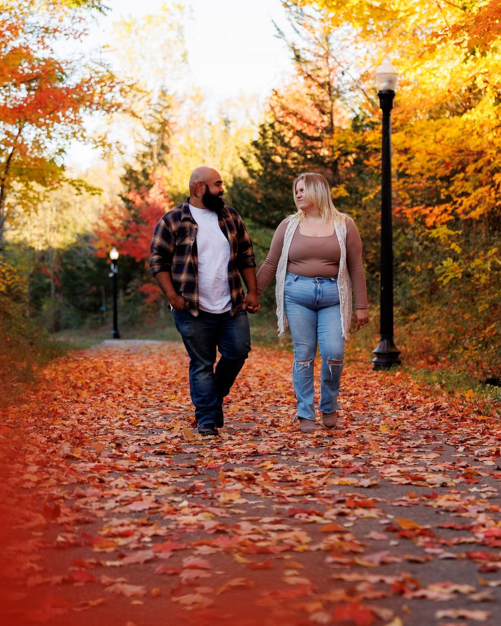 A couple walks along a path covered in vibrant autumn leaves with golden trees in the background during sunset.