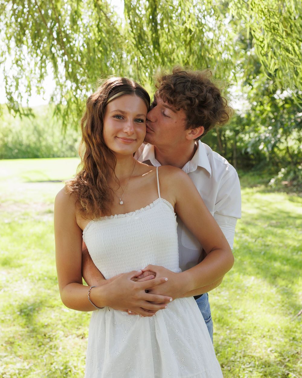 A romantic couple embraces under a weeping willow tree on a sunny summer day.